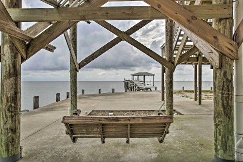 A wooden swing hangs under a structure, overlooking a calm waterfront with a cloudy sky and distant pier.