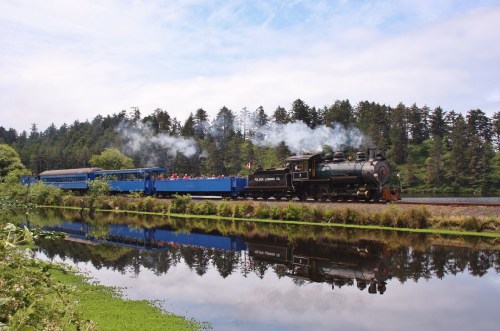 A steam train travels along a scenic route beside a reflective body of water, surrounded by lush greenery and trees.