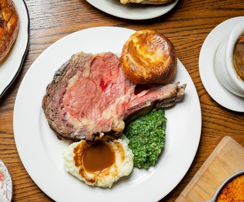 A plate featuring a large slice of prime rib, mashed potatoes with gravy, green sauce, and a Yorkshire pudding.