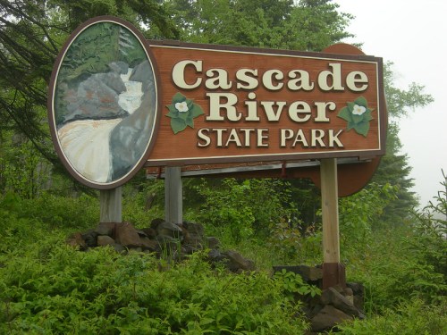 Wooden sign for Cascade River State Park, featuring a waterfall illustration and surrounded by greenery.