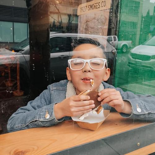 A child with glasses enjoys a s'more, smiling while sitting by a window at a café.