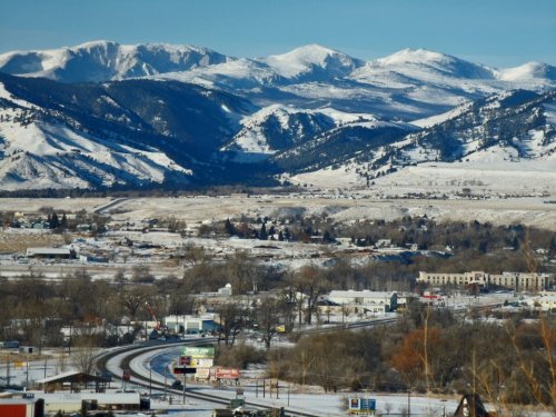 Snow-covered mountains rise in the background, with a valley and small town visible in the foreground under clear blue skies.
