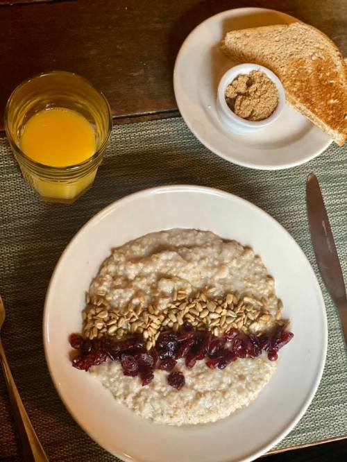 A bowl of oatmeal topped with cranberries and sunflower seeds, served with toast, brown sugar, and orange juice.
