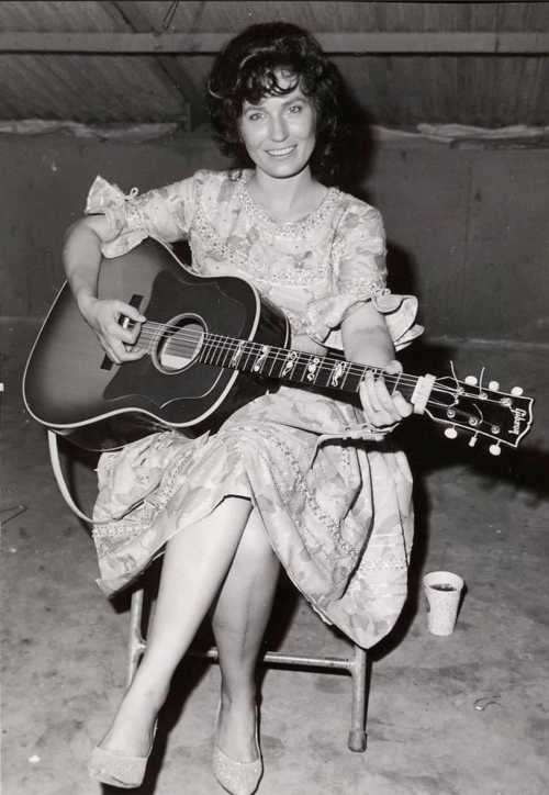 A woman in a vintage dress sits on a chair, playing an acoustic guitar and smiling at the camera.