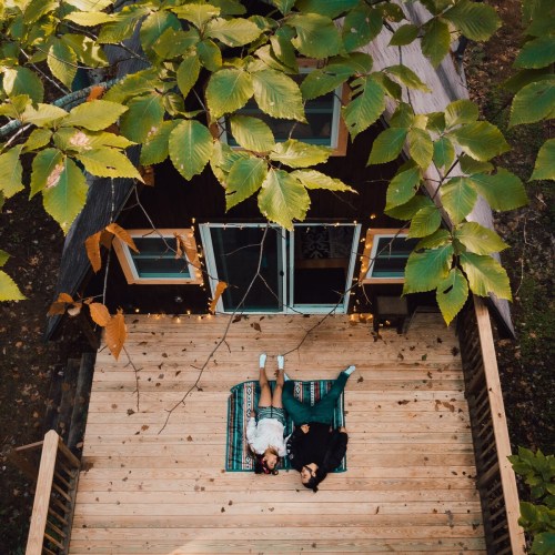 Two people relax on a wooden deck surrounded by greenery, lying on a blanket under a canopy of leaves.