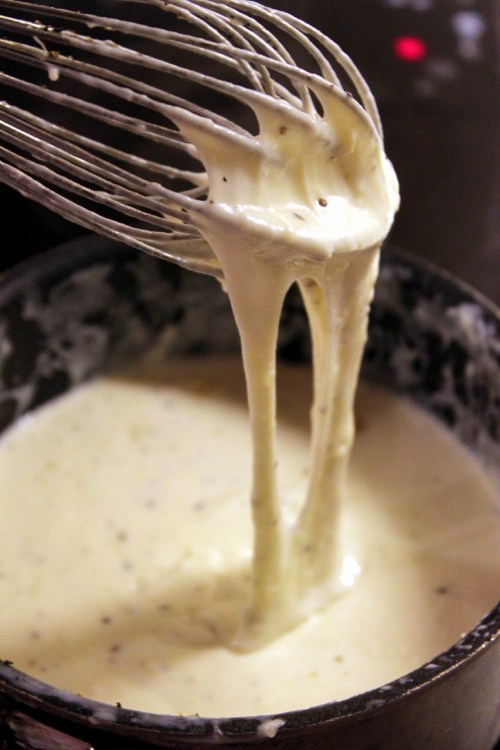 A whisk lifting creamy, thick sauce from a pot, with strands of sauce stretching between the whisk and the pot.
