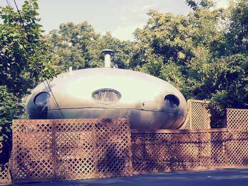 A futuristic, metallic pod-shaped structure surrounded by wooden lattice fencing and greenery.