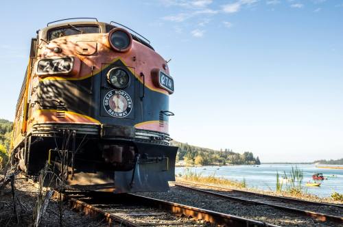 A vintage train on tracks by a river, surrounded by trees and hills, with a clear blue sky in the background.