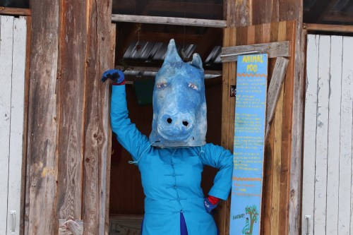A person in a blue horse mask poses in a barn doorway, wearing a blue outfit and colorful gloves.