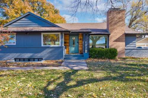 A blue single-story house with a chimney, front porch, and autumn foliage in the yard.