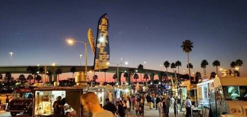 A bustling food truck event at dusk, with palm trees and a colorful sky in the background.