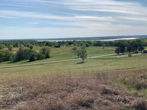 A scenic view of a green landscape with trees, a winding road, and a lake in the distance under a blue sky.