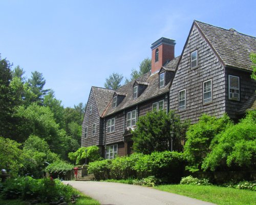 A large, rustic house with a shingled roof surrounded by lush greenery and trees under a clear blue sky.