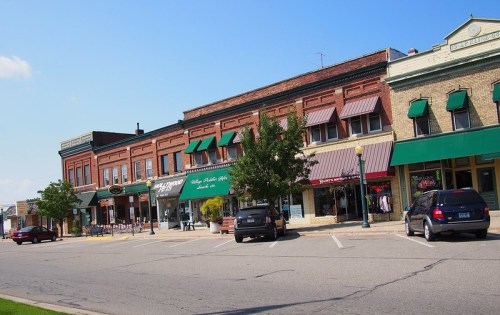 A charming small-town street featuring brick buildings with shops, awnings, and parked cars under a clear blue sky.