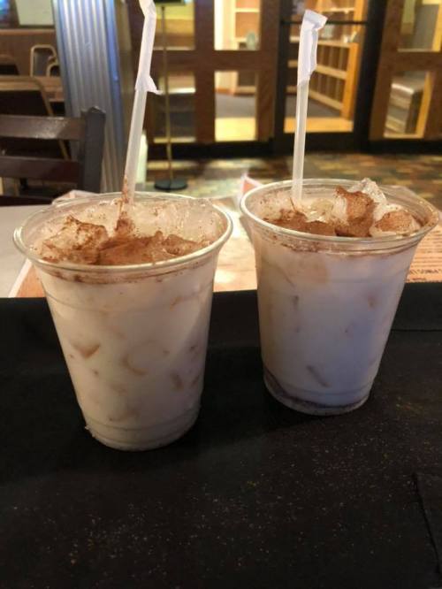 Two clear cups filled with a creamy beverage and ice, topped with a sprinkle of cinnamon, sit on a table.