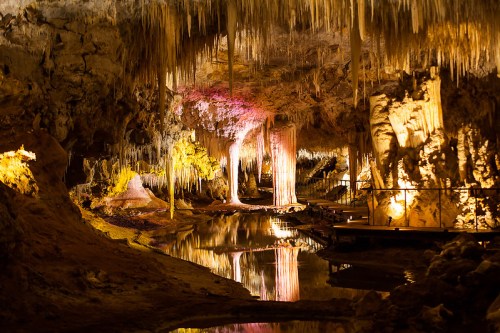 Stalactites and stalagmites in a dimly lit cave, reflecting in a calm pool of water. Colorful lights enhance the scene.