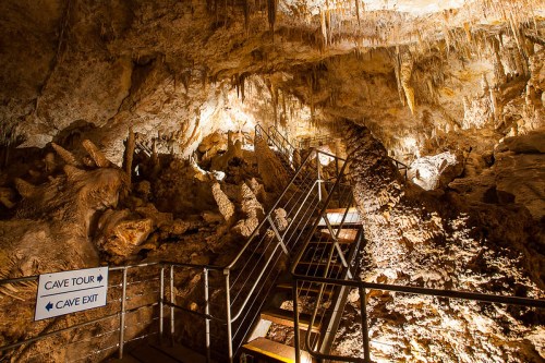 A dimly lit cave interior with rock formations and a metal staircase leading through the cave tour area.