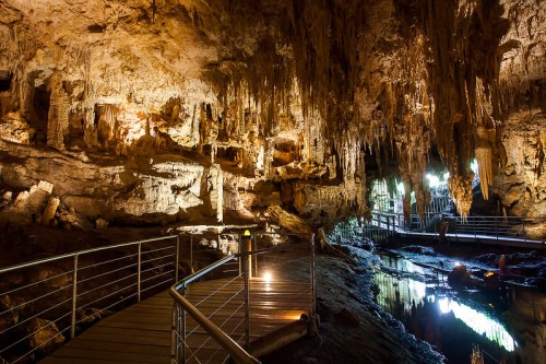 A dimly lit cave interior with stalactites, a wooden walkway, and reflections in a calm water pool.