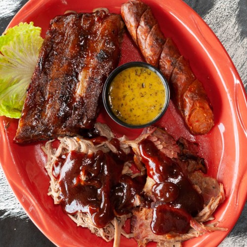 A red plate with BBQ ribs, sausage, pulled pork with sauce, and a side of mustard sauce, garnished with lettuce.