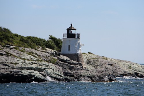 A white lighthouse stands on rocky shore, surrounded by greenery and ocean under a clear blue sky.