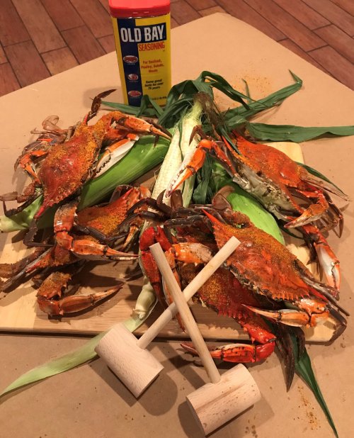 A wooden board with seasoned crabs, corn, and two mallets, alongside a container of Old Bay seasoning.