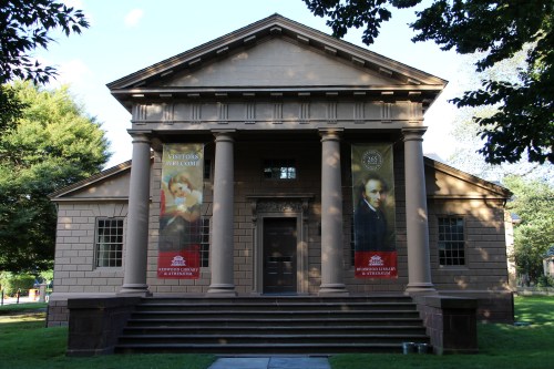 Historic building with columns, featuring banners on either side, surrounded by greenery and a clear blue sky.