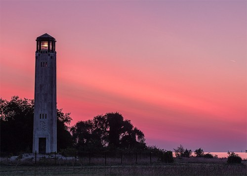 A tall lighthouse silhouette against a vibrant pink and purple sunset sky, surrounded by trees and grass.
