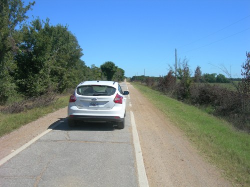 A white car parked on a rural road with trees and a clear blue sky in the background.
