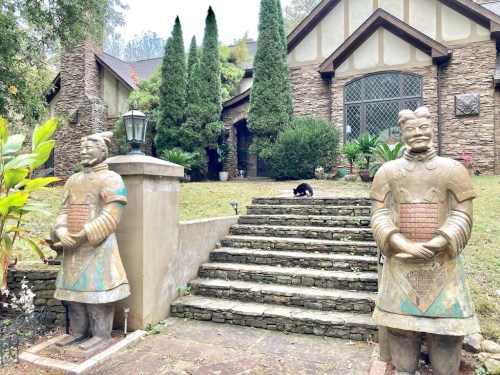Two large statues flank stone steps leading to a house, with a black cat sitting on the grass in front.