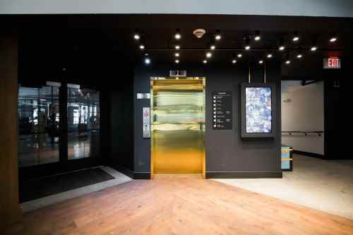 A modern elevator with a gold door in a stylish lobby, featuring dim lighting and a digital directory nearby.