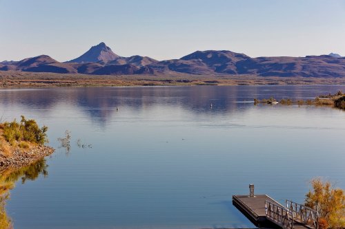 A calm lake reflects mountains under a clear sky, with a small dock visible in the foreground.