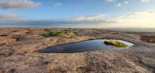 A serene landscape featuring a small pond surrounded by rocky terrain and sparse greenery under a cloudy sky.