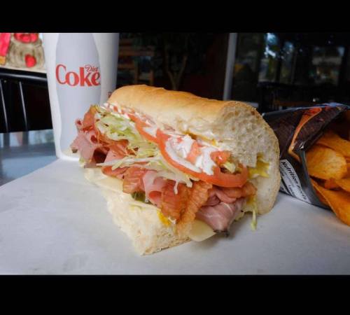 A sub sandwich filled with meats, lettuce, tomatoes, and mayonnaise, alongside a Diet Coke and a bag of chips.
