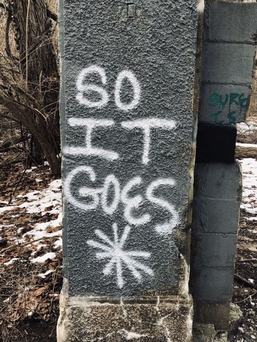 Graffiti on a stone pillar reads "SO IT GOES" in white spray paint, surrounded by bare trees and snow.
