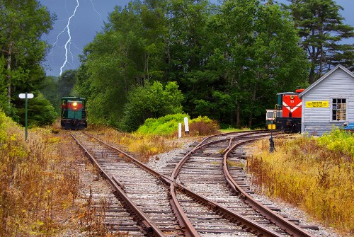 A stormy scene with lightning, featuring two trains on diverging tracks near a small station surrounded by trees.
