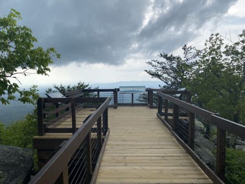 A wooden observation deck surrounded by trees, overlooking a cloudy sky and distant hills.