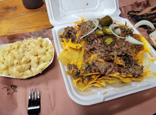 A takeout container with nachos topped with beef, jalapeños, and onions, alongside a side of macaroni and cheese.