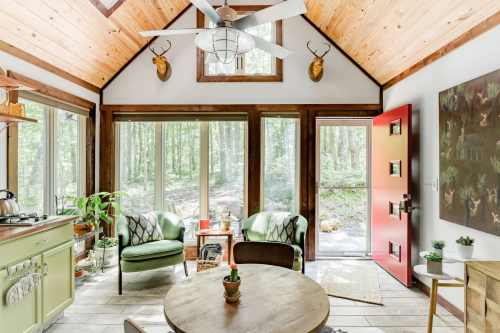 Cozy cabin interior with wooden beams, green accents, and large windows overlooking a forest. Red door adds a pop of color.