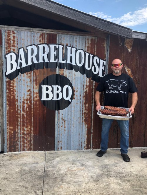 A man in sunglasses holds a tray of ribs in front of a rustic BBQ restaurant sign that reads "Barrelhouse BBQ."