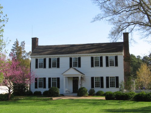 A classic white two-story house with black shutters, surrounded by green grass and blooming trees.