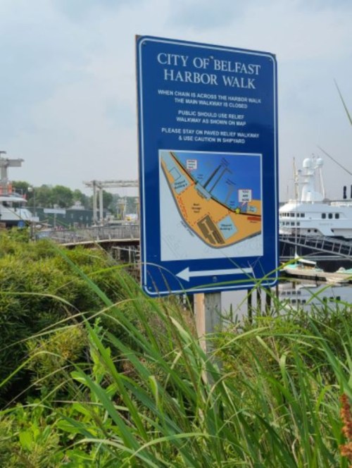 Sign for the City of Belfast Harbor Walk, detailing access and safety information, surrounded by greenery.