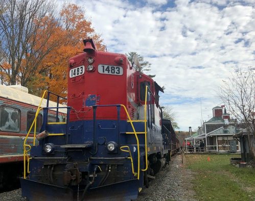 A red and blue train engine parked on tracks, surrounded by autumn trees and a cloudy sky.
