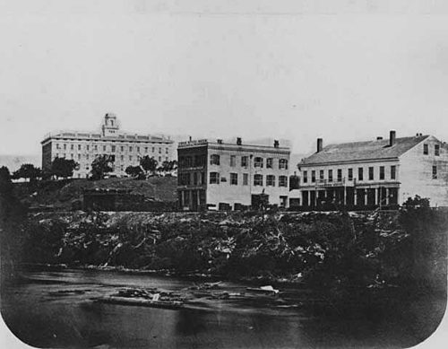 Historic black-and-white photo of three buildings along a riverbank, surrounded by trees and a hilly landscape.
