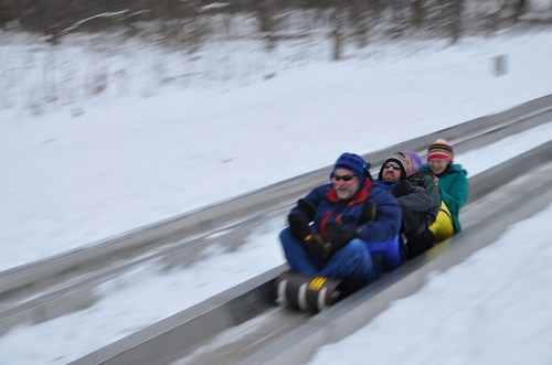 A group of four people joyfully sledding down a snowy slope, surrounded by winter scenery.