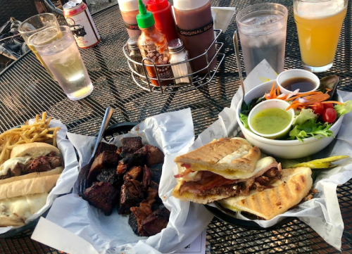 A table with various dishes: sandwiches, fries, grilled meat, salad, and drinks, set outdoors.