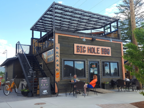 A two-story BBQ restaurant with outdoor seating, a staircase, and a bike parked outside on a sunny day.