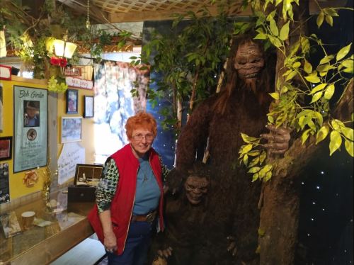 A woman stands next to a large Bigfoot statue in a cozy, decorated room filled with memorabilia.