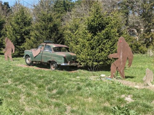 An old green car surrounded by wooden Bigfoot cutouts in a grassy area with trees in the background.