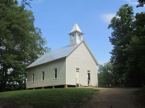 A small white church with a steeple, surrounded by trees and a gravel path under a clear blue sky.