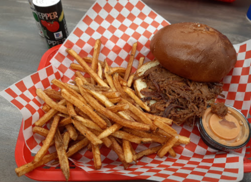 A plate with a pulled pork sandwich on a bun, crispy fries, and a small cup of dipping sauce, alongside a soda can.